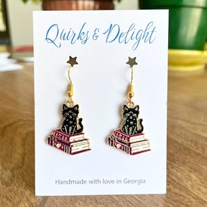 May include: Gold earrings featuring a black cat sitting on a stack of three books with a purple and gold design. The earrings are on a white card with the text "Quirks & Delight" and "Handmade with love in Georgia".