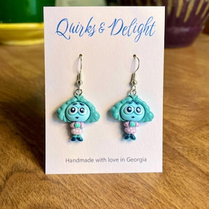 Könnte beinhalten: Ein Paar handgefertigte Ohrringe mit einem Cartoon-Charakter-Design. Die Ohrringe haben blaue Haare, große Augen und tragen ein rosa Kleid mit weißen Punkten. Auf der Karte steht "Quirks & Delight" und "Handmade with love in Georgia."