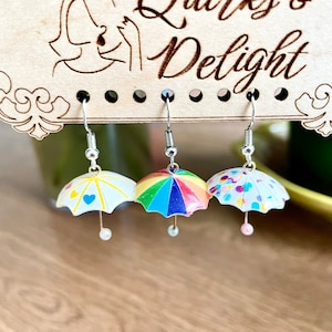 Könnte beinhalten: Drei Paar Ohrringe mit Miniatur-Regenschirmen. Die Regenschirme sind mit verschiedenen Mustern verziert: einer mit gelben und blauen Herzen, einer mit einem Regenbogen und einer mit bunten Konfetti.