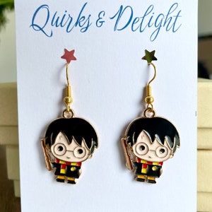 May include: A pair of gold-colored dangle earrings featuring a cartoon character with black hair, glasses, and a wand. The earrings are on a white card with the text "Quirks & Delight" and "Handmade with love in Georgia".