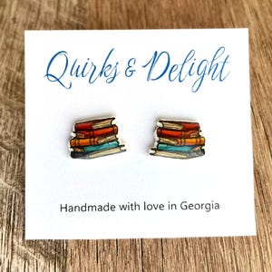 Op de afbeelding: Stud oorbellen in de vorm van gestapelde boeken. De boeken hebben rode, oranje en turquoise ruggen. De oorbellen worden gepresenteerd op een witte kaart met de tekst "Quirks & Delight" en "Handmade with love in Georgia".