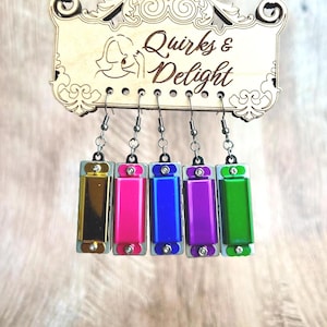 May include: Five pairs of earrings featuring miniature harmonicas in different colors: gold, pink, blue, purple, and green. The earrings are hanging from a wooden sign that reads "Quirks & Delight."