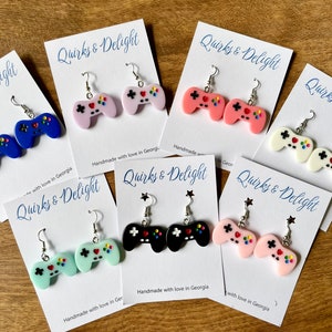 Könnte beinhalten: Ohrringe mit bunten Videospiel-Controller-Designs in Blau, Rosa, Schwarz, Weiß und Mintgrün. Jedes Paar wird auf einer weißen Karte mit dem Text "Quirks & Delight" und "Handmade with love in Georgia" präsentiert.