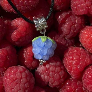 May include: A pendant shaped like a blue berry with green leaves at the top, suspended from a black cord. The berry has a translucent, textured surface and is accented with a small silver-toned metal bail and a tiny silver-toned bead at the base.