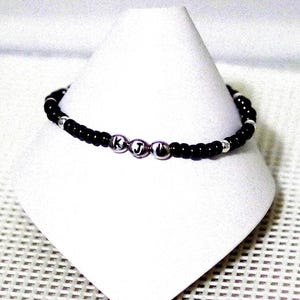 May include: A black beaded bracelet with silver beads that spell out the letters "K J" in a simple font.