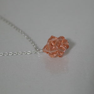 Puede incluir: Un collar de cadena de plata con un colgante de cristal de color melocot&oacute;n. El colgante es una forma geom&eacute;trica con muchas facetas.