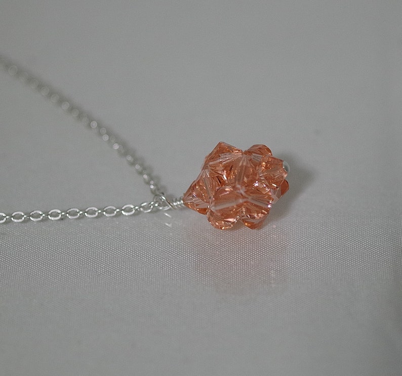 Puede incluir: Un collar de cadena de plata con un colgante de cristal de color melocot&oacute;n. El colgante es una forma geom&eacute;trica con muchas facetas.