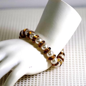 May include: A bracelet made of white pearls and brown beads. The bracelet is on a white mannequin hand.