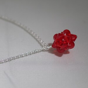 Puede incluir: Un collar de cadena de plata con un colgante de cristal rojo. El colgante es una forma geom&eacute;trica con muchas facetas.