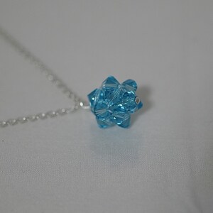 Puede incluir: Un collar de cadena de plata con un colgante de cristal azul claro. El colgante tiene forma de estrella con muchas facetas.