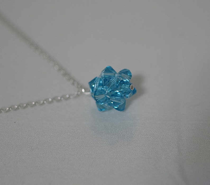 Puede incluir: Un collar de cadena de plata con un colgante de cristal azul claro. El colgante tiene forma de estrella con muchas facetas.