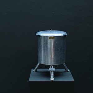 May include: A cylindrical silver-colored metal container with a flat lid, supported by three sturdy legs. A foot-operated pedal mechanism is positioned between the front legs. A small rectangular label with the text ADORA is attached to the front.