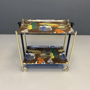 Op de afbeelding: Een serveerwagen met twee niveaus en een bruin oppervlak met een kleurrijk patroon van fruit en kannen. De wagen heeft een goudkleurig metalen frame, zwarte verticale steunen en vier kleine wieltjes aan de basis. De bladen hebben opstaande randen.
