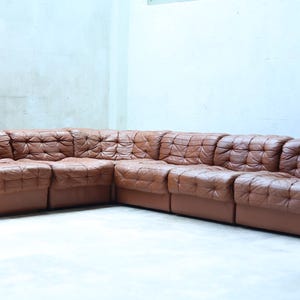 Op de afbeelding: Een grote, bruine leren hoekbank met een gecapitonneerd ontwerp. De bank staat in een hoekopstelling en lijkt vintage te zijn. Het leer heeft een rijke, warme tint en de kussens zijn vierkant.