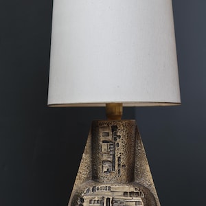 Op de afbeelding: Een vintage tafellamp met een witte lampenkap en een bronskleurige voet. De voet heeft een geometrisch ontwerp met architecturale details. De lamp staat op een grijs oppervlak tegen een donkergrijze achtergrond.