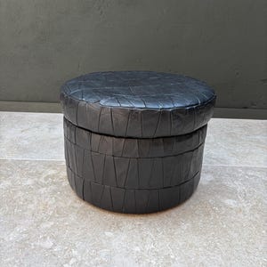 Op de afbeelding: Een ronde zwarte poef opgebouwd uit meerdere gestikte lederen panelen. Het meubelstuk heeft een cilindrische basis en een platte ronde bovenkant, beide bekleed met een patchwork van donkere lederen segmenten in een geometrisch patroon.