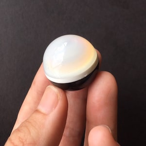 May include: A spherical, decorative object with a glossy finish. The top half is white, transitioning to a yellow hue, with a black band at the base. The object is held between fingers against a dark background.