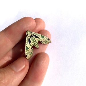 Könnte beinhalten: Eine grüne und schwarze Emaille-Anstecknadel in Form eines Nachtfalters mit detaillierten Flügeln.