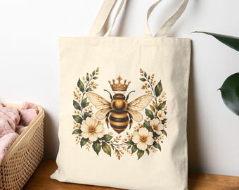 May include: A cream-coloured canvas tote bag featuring a central graphic of a bee wearing a small crown, surrounded by a circular wreath of white flowers and green leaves. The bag has two long shoulder straps and a rectangular body shape.