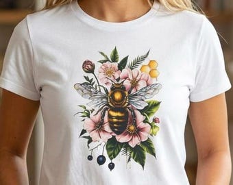 May include: A white short-sleeved t-shirt featuring a central graphic print of a detailed honeybee surrounded by pink flowers, green leaves, dark berries, and yellow honeycomb shapes. The shirt is paired with dark blue denim jeans.