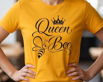 May include: A mustard yellow short-sleeved t-shirt featuring a black graphic print on the chest. The design includes a stylised crown above the cursive text "Queen Bee" and a small bee illustration with striped wings positioned below the text.
