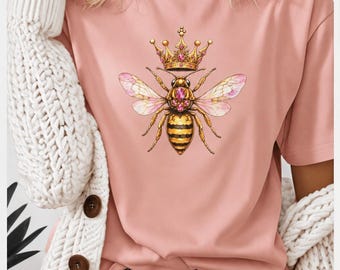 May include: A dusty pink short-sleeved t-shirt featuring a central graphic of a bee wearing a gold crown with pink gemstones. The bee has detailed wings with pink accents and a striped body. The shirt is worn with a white cable-knit cardigan.