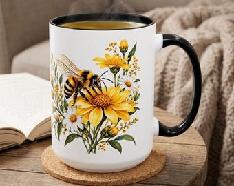 May include: A white ceramic mug with a black rim and handle, featuring a graphic illustration of a honeybee resting on a large yellow flower surrounded by smaller white daisies, green leaves, and yellow buds, with steam rising from the top.