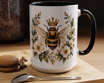 May include: A white ceramic mug with a black interior and handle, featuring a central illustration of a bee wearing a gold crown, surrounded by a wreath of white flowers and green leaves. The design is printed on the front of the mug.