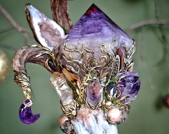 May include: A handcrafted, ornate staff featuring a large, faceted purple crystal at the top. The staff is adorned with various crystals, gold-colored metalwork, and a small purple crescent moon charm. The design incorporates natural elements like antlers and feathers.
