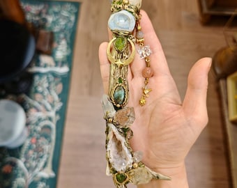 May include: A handcrafted wand featuring clear quartz crystals, a light blue stone, and various green and clear gemstones. The wand has a gold-colored metal frame with decorative accents and a dangling charm. The wand is approximately 12 inches long.