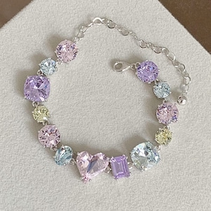 Peut inclure: Un bracelet en argent avec un pendentif en forme de cœur et des pierres précieuses colorées. Le bracelet présente une variété de pierres de couleur pastel, notamment rose, violet et jaune.