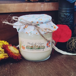 Peut inclure: Un pot en verre rempli de cire blanche, surmonté d'un couvercle en tissu blanc à motifs floraux orange et verts, noué avec une ficelle de jute naturelle. L'étiquette porte le texte Pêche de Vigne à la douce lumière avec des illustrations de pêches.