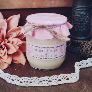 Peut inclure: Un pot en verre rempli d'une substance crémeuse, surmonté d'un couvercle en tissu rose à motifs floraux blancs, noué par une ficelle brune. L'étiquette porte l'inscription BARBE À PAPA et A LA DOUCE LUMIÈRE, entourée de dessins de barbe à papa.