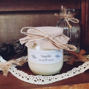 Peut inclure: Une bougie en verre dans un bocal avec un couvercle en toile de jute et une étiquette qui indique "Vanille & A la douce lumière". La bougie est posée sur une surface en bois avec une dentelle et d'autres objets décoratifs.