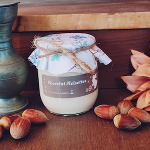 Peut inclure: Un pot en verre avec un couvercle blanc et un nœud en ficelle brune. Le pot est rempli d'une tartinade brun clair et porte une étiquette sur laquelle est inscrit "Chocolat Noisettes" et "Au doux lumière de..." Le pot est posé sur une table en bois avec des noisettes et un pichet en métal.