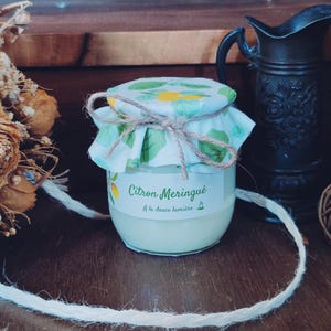 Peut inclure: Un petit pot en verre de dessert citron meringué, étiqueté "Citron Meringué". Le pot est recouvert d'un couvercle en tissu noué avec de la ficelle. Des fleurs séchées et une cruche décorative sont en arrière-plan.