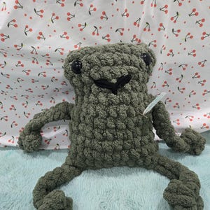 May include: A handmade, olive green crochet frog plush toy with black button eyes and a stitched black mouth. The frog has outstretched arms and legs. It is displayed on a light blue surface with a cherry print background.