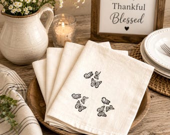 May include: A set of off-white cloth napkins featuring a black printed butterfly pattern. The napkins are folded and stacked inside a round wooden bowl, displaying a repeating design of various butterfly shapes across the fabric surface.