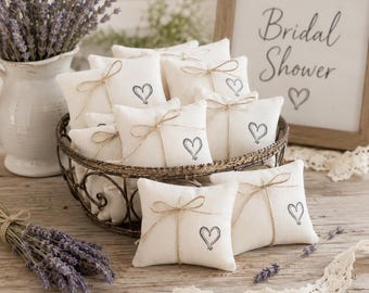 May include: A collection of small, square, off-white fabric sachets filled with lavender, each tied with thin twine and featuring a printed heart design. The sachets are arranged in a dark metal wire basket, with a sign reading Bridal Shower.