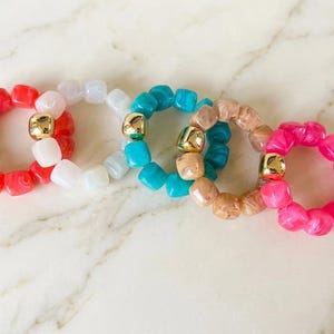 Peut inclure: Quatre bagues circulaires composées de petites perles carrées rouges, blanches, turquoise et roses, chacune dotée d'une perle métallique dorée, lisse et réfléchissante. Les perles ont une finition marbrée semi-translucide et sont enfilées.