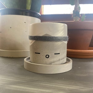 May include: A small, white concrete planter with a gray rope band around the middle. The planter has a simple, hand-painted face with two black dashes for eyes and a black circle for a mouth.
