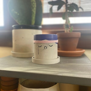 May include: A small, white ceramic planter with a blue rim and a pink band around the top. The planter has a sad face drawn on it with a single tear.