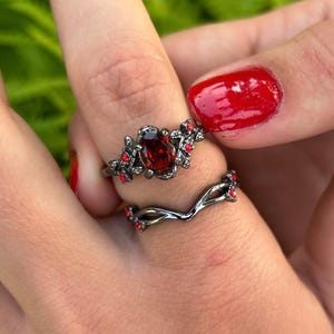 Op de afbeelding: Twee ringen worden op een vinger getoond. De bovenste ring heeft een donkere metalen band met een grote, ovale, rode edelsteen en kleinere rode accenten. De tweede ring is een gebogen band met rode accenten. De ringen staan tegen een wazige groene achtergrond.