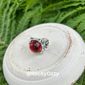 Puede incluir: Un anillo plateado con una gema roja grande y redonda. El anillo tiene un diseño detallado y texturizado alrededor de la gema, y la gema tiene un patrón oscuro y arremolinado. El anillo está sobre una superficie blanca con follaje verde en el fondo.