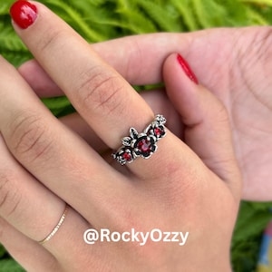 Peut inclure: Bague argentée ornée d'un motif floral avec trois pierres rouges. La bague présente un aspect détaillé et texturé, les pierres étant serties au centre de chaque fleur. La bague est portée au doigt.