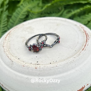 Puede incluir: Dos anillos negros con detalles rojos. Un anillo presenta una gran gema roja ovalada rodeada de un diseño floral. El otro anillo tiene forma de V con pequeños detalles rojos. Los anillos se muestran sobre una superficie blanca texturizada.