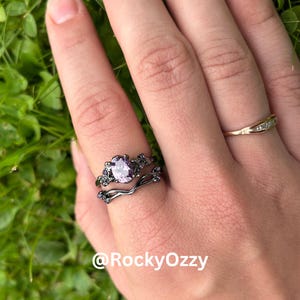 Può includere: Una mano mostra tre anelli: un anello di fidanzamento nero con una pietra centrale viola chiaro e dettagli floreali, una fascia nera abbinata e una fascia dorata con piccole pietre trasparenti. Gli anelli sono su uno sfondo verde.