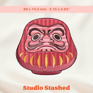 May include: A red Daruma doll embroidery design, measuring 80 x 74.5 mm. The doll has a stern expression with dark brown eyebrows and eyes. Gold and yellow accents are visible on the lower portion of the doll. The text "Studio Stashed" is at the bottom.