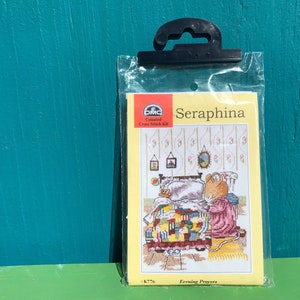 May include: A DMC counted cross stitch kit featuring a mouse named Seraphina saying her evening prayers. The kit includes a colorful image of Seraphina kneeling beside her bed with a patchwork quilt. The kit is titled "Seraphina: Evening Prayers" and has the DMC logo.