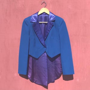 May include: A blue tailcoat jacket with a purple satin lining and lapels, hanging on a wooden hanger. The jacket has a single button closure and long sleeves. The background is a textured, reddish-pink surface.